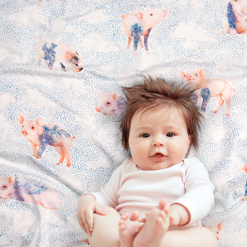 
                      
                        Baby resting on Pima cotton blanket with japanese inspired watercolor artwork showing a variety of tattooed pigs, on a background of tiny dots and clouds.
                      
                    